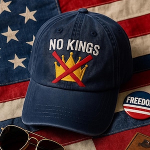 Puede incluir: Gorra de béisbol azul marino con el texto bordado "NO KINGS" sobre una corona dorada tachada. Un botón "FREEDOM" rojo, blanco y azul y gafas de sol con montura dorada descansan sobre una bandera americana.