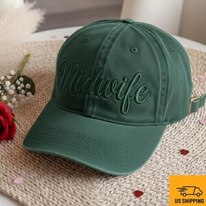 May include: A dark green baseball cap with the word "Midwife" embroidered in a matching tone. The cap has a curved brim and an adjustable strap. The hat is sitting on a woven surface with a red rose and small heart-shaped decorations.