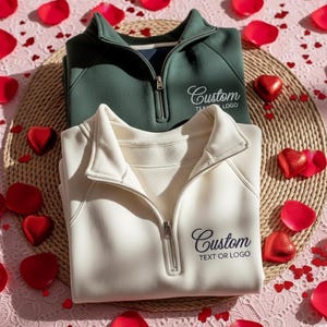 Puede incluir: Dos jerséis con cremallera de un cuarto, uno crema y otro verde oscuro, se exhiben sobre una esterilla tejida. Cada uno tiene un logotipo personalizado bordado en el pecho. Pétalos de rosa rojos y bombones en forma de corazón rodean los jerséis sobre una superficie rosa.