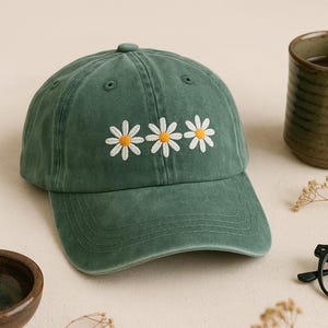 May include: A green baseball cap with three embroidered daisy flowers. The cap has a curved brim and a button on top. The daisies have white petals and yellow centers. The cap is made of a soft, worn material.