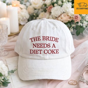 May include: White baseball cap with the embroidered text "THE BRIDE NEEDS A DIET COKE" in red. The cap has a curved brim and a classic six-panel design. Perfect for a bachelorette party or wedding.