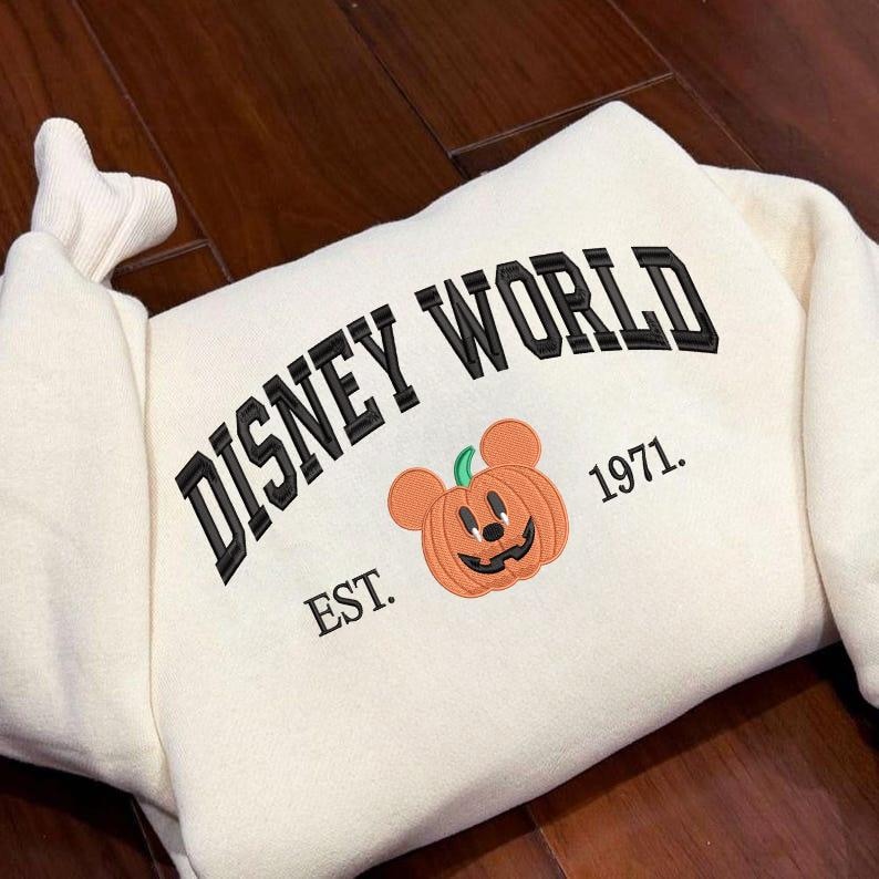 Może przedstawiać: Kremowa bluza z haftowanym czarnym napisem "DISNEY WORLD". Poniżej znajduje się uśmiechnięta dynia z uszami Myszki Miki. Bluza ma r&oacute;wnież napis "EST. 1971."