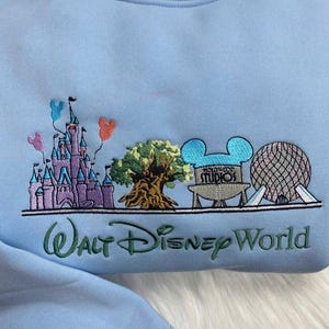 Może przedstawiać: Jasnoniebieska bluza z haftowanym wzorem przedstawiającym zamek, drzewo, logo Hollywood Studios i kulę geodezyjną. Pod spodem wyhaftowano napis "Walt Disney World". W zestawie znajdują się r&oacute;wnież balony z Myszki Miki.