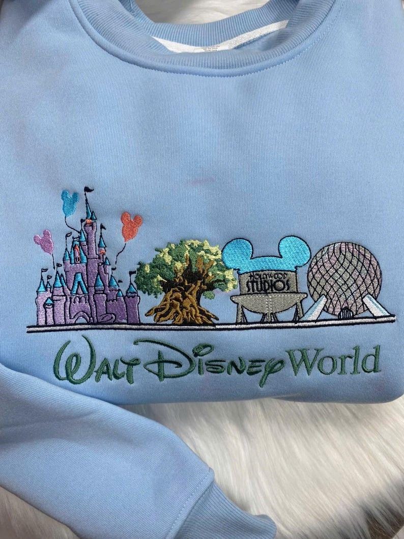 Może przedstawiać: Jasnoniebieska bluza z haftowanym wzorem przedstawiającym zamek, drzewo, logo Hollywood Studios i kulę geodezyjną. Pod spodem wyhaftowano napis "Walt Disney World". W zestawie znajdują się r&oacute;wnież balony z Myszki Miki.