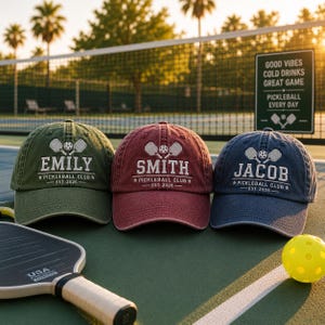 K&ouml;nnte beinhalten: Drei personalisierte Baseballkappen in Olivgr&uuml;n, Burgunderrot und Marineblau, jede bestickt mit einem Namen, Pickleballschl&auml;gern und dem Text "Pickleball Club - Est. 2026." Ein Pickleballschl&auml;ger und -ball befinden sich im Vordergrund.