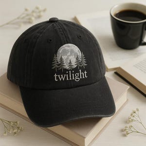 Może przedstawiać: Czarna czapka bejsbolówka z haftowanym wzorem przedstawiającym pełnię księżyca, sosny i słowo "twilight" w kolorze białym. Czapka leży na książkach, z filiżanką kawy w tle.
