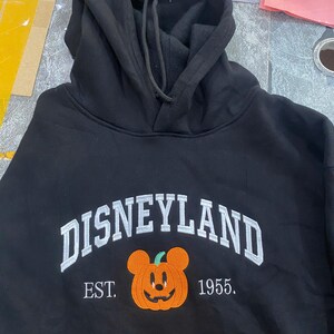 Może przedstawiać: Czarna bluza z kapturem z białym haftem "DISNEYLAND", uśmiechniętą pomarańczową dynią z uszami Myszki Miki i haftowanym tekstem "EST. 1955."