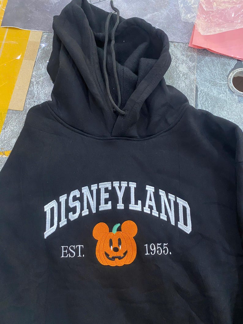 Może przedstawiać: Czarna bluza z kapturem z białym haftem "DISNEYLAND", uśmiechniętą pomarańczową dynią z uszami Myszki Miki i haftowanym tekstem "EST. 1955."