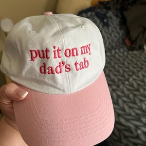 Puede incluir: Gorra de b&eacute;isbol blanca y rosa con la frase bordada "put it on my dad's tab" en rojo. La gorra tiene una visera rosa y un bot&oacute;n rosa en la parte superior. El sombrero est&aacute; hecho de tela suave.