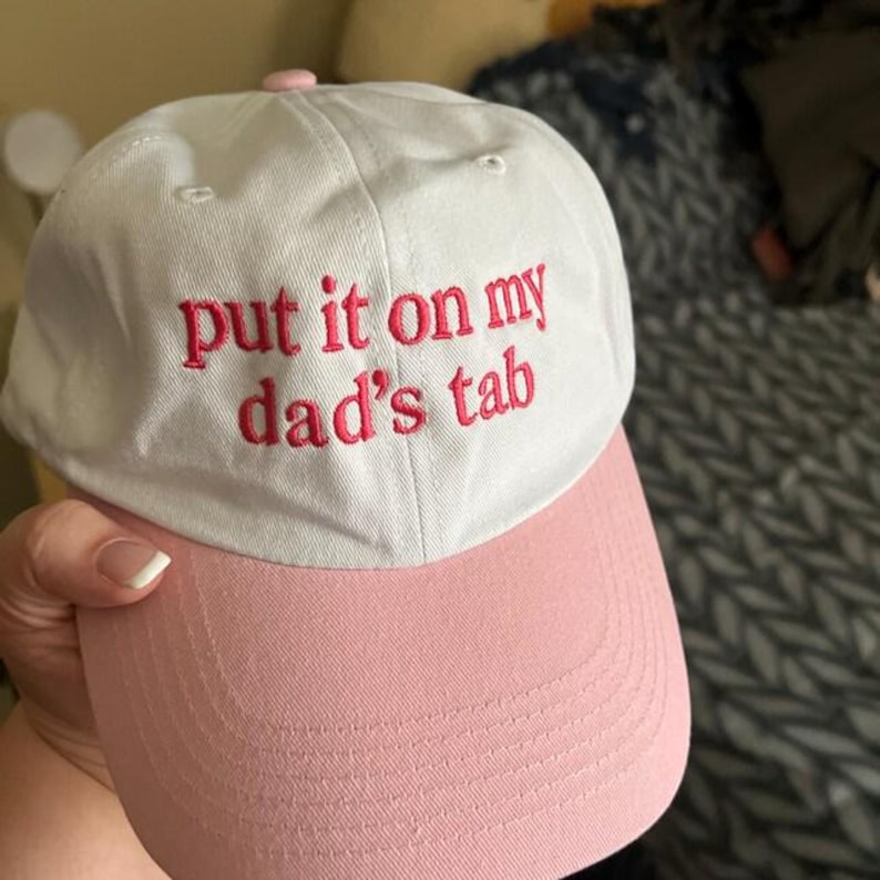 Puede incluir: Gorra de b&eacute;isbol blanca y rosa con la frase bordada "put it on my dad's tab" en rojo. La gorra tiene una visera rosa y un bot&oacute;n rosa en la parte superior. El sombrero est&aacute; hecho de tela suave.