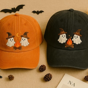 May include: Two baseball caps, one orange and one black, each with an embroidered Halloween design. The design features two ghosts wearing witch hats, a campfire, and a crescent moon. The caps are surrounded by pumpkins, bats, and pinecones.