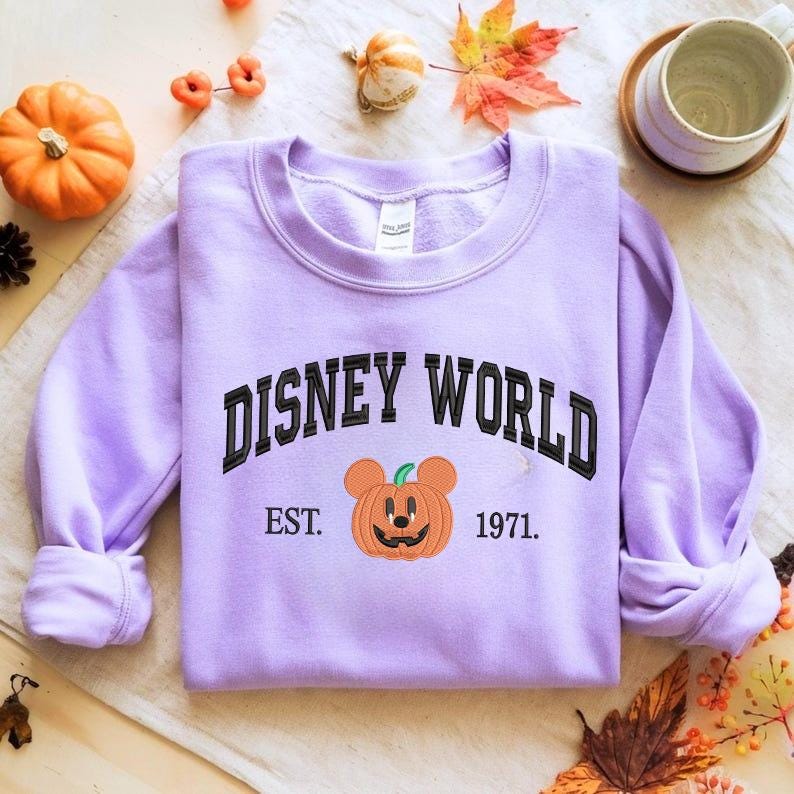 Może przedstawiać: Jasnofioletowa bluza z napisem "DISNEY WORLD" czarnymi literami. Poniżej haftowana dynia z uszami Myszki Miki. Pod dynią znajduje się napis "EST. 1971".