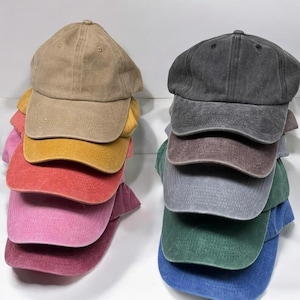 May include: Stacks of baseball caps in various colors, including tan, yellow, red, pink, gray, brown, green, and blue. Each cap has a curved brim and a button on top. The caps are made of a soft, washed fabric.