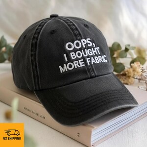 Peut inclure: Casquette de baseball noire avec le texte blanc brodé "OOPS, I BOUGHT MORE FABRIC". Le chapeau est posé sur un livre. La casquette a une visière incurvée et une sangle réglable.
