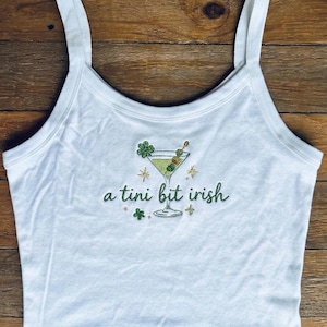 May include: White spaghetti strap tank top with green embroidered martini glass design and the phrase "a tini bit irish". The martini has an olive and shamrock garnish. The design is surrounded by star and shamrock accents.