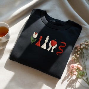 May include: Black sweatshirt with embroidered design featuring a white tulip, a red chess piece, a white chess piece, a red apple held by white hands, and a red ribbon. The sweatshirt is displayed on a light-colored fabric with a cup of tea and flowers.