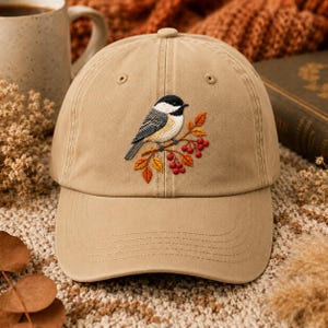 Puede incluir: Una gorra de béisbol de color tostado con un bordado de un carbonero cabecinegro posado en una rama con hojas de colores otoñales y pequeñas bayas rojas. La gorra presenta una corona estructurada, una visera curva y ojales cosidos.