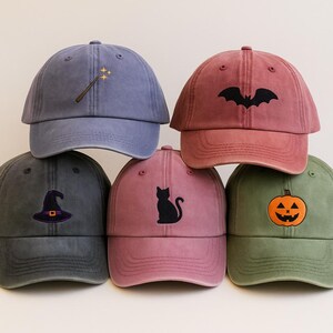 May include: A selection of baseball caps in different colours, each with an embroidered Halloween design. Designs include a witch's hat, a black cat, a jack-o'-lantern, a bat, and a magic wand. The caps are made from a soft, washed fabric.