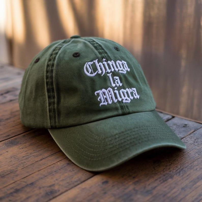 K&ouml;nnte beinhalten: Olivgr&uuml;ne Baseballkappe mit dem gestickten Text "Chinga La Migra" in Wei&szlig;. Die Kappe hat einen gebogenen Schirm und ein klassisches Sechs-Panel-Design. Der Hut ist aus strapazierf&auml;higem Stoff gefertigt und liegt auf einer Holzoberfl&auml;che.
