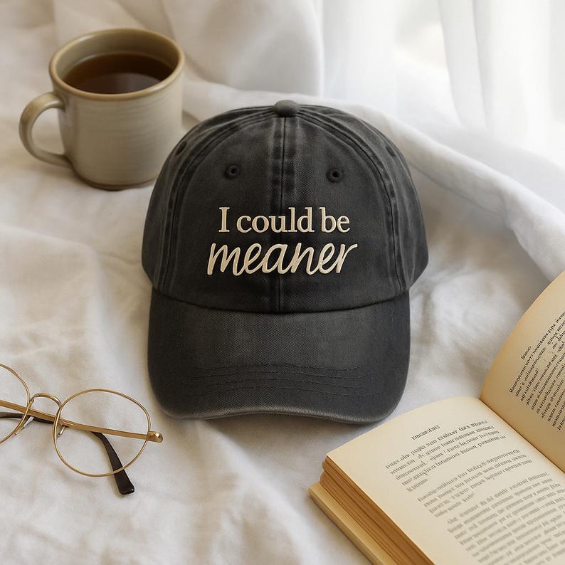 May include: A dark gray baseball cap with the embroidered text "I could be meaner" in white. The cap is slightly worn, with a curved brim. A beige mug and a pair of gold-rimmed glasses are also visible.