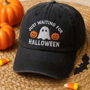 Peut inclure: Casquette de baseball noire avec le texte brodé "JUST WAITING FOR HALLOWEEN". Le motif comprend un fantôme blanc et deux citrouilles orange. La casquette est sur une surface tissée avec des citrouilles et des bonbons.