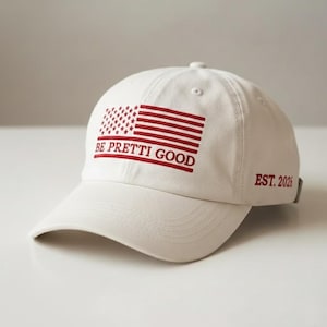 Puede incluir: Gorra de béisbol blanca con visera curva. La gorra presenta un diseño de bandera americana bordada en rojo y el texto "BE PRETTI GOOD". El texto "EST. 2026" también está bordado en el lateral.