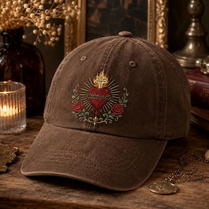 Può includere: Un cappellino da baseball marrone con un ricamo di un sacro cuore rosso sormontato da una croce dorata e fiamme, circondato da tralci di foglie verdi e due rose rosse. Il cappellino presenta una corona strutturata e una visiera curva.