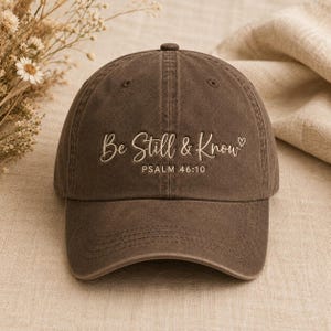 Könnte beinhalten: Eine dunkelbraune Baseballkappe mit verwaschener Optik. Auf der Vorderseite befindet sich eine hellbeige Stickerei mit dem Text "Be Still & Know" in Schreibschrift, gefolgt von "PSALM 46:10" in serifenloser Schrift und einem kleinen Herz.
