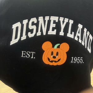 Może przedstawiać: Czarna bluza z białym haftem "Disneyland" w łuku. Poniżej haftowana pomarańczowa dynia z uszami Myszki Miki. Na bluzie wyhaftowano r&oacute;wnież napis "EST. 1955.".