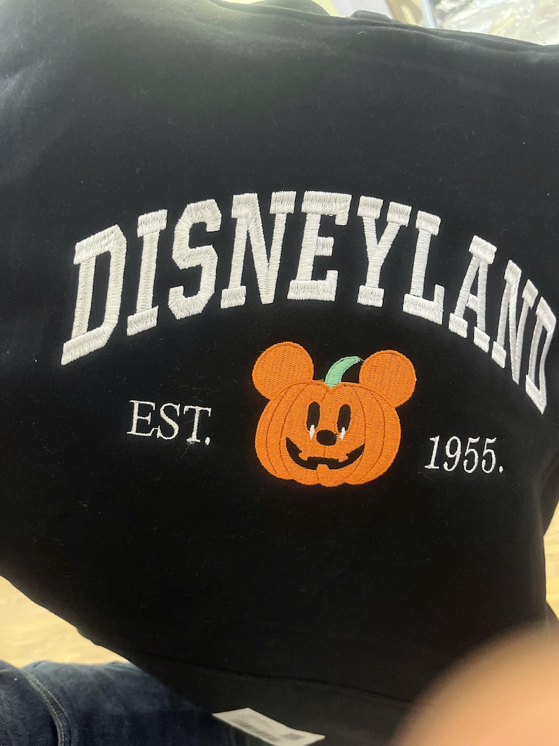 Może przedstawiać: Czarna bluza z białym haftem "Disneyland" w łuku. Poniżej haftowana pomarańczowa dynia z uszami Myszki Miki. Na bluzie wyhaftowano r&oacute;wnież napis "EST. 1955.".