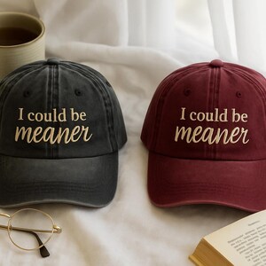 May include: Two baseball caps, one dark gray and one burgundy, each with the embroidered text "I could be meaner." A pair of round eyeglasses and a mug of tea are also visible.