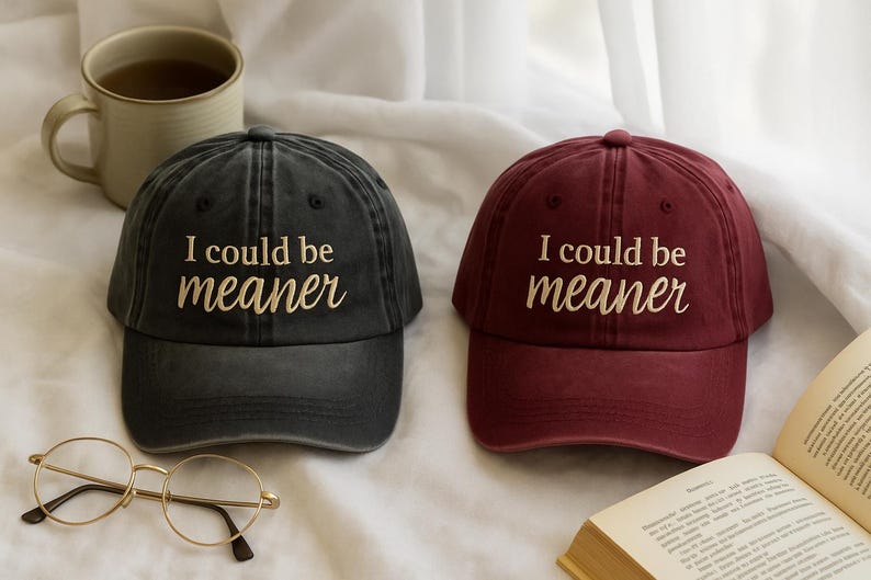 May include: Two baseball caps, one dark gray and one burgundy, each with the embroidered text "I could be meaner." A pair of round eyeglasses and a mug of tea are also visible.