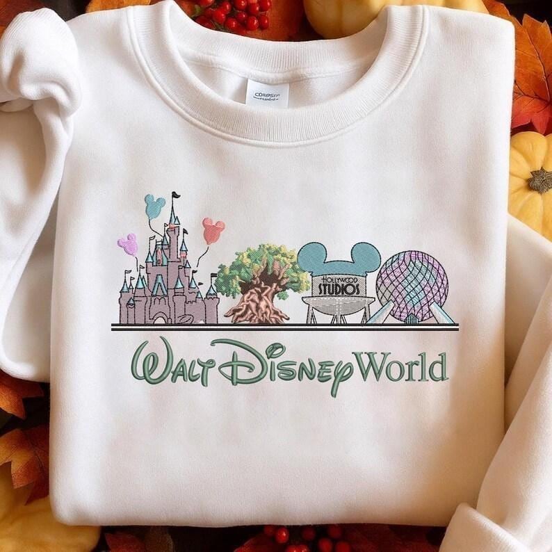 Może przedstawiać: Biała bluza z haftowanym wzorem ikon Disney World, w tym zamku, drzewa i Spaceship Earth. Tekst "Walt Disney World" jest w kolorze zielonym poniżej. Bluza ma okrągły dekolt i długie rękawy.