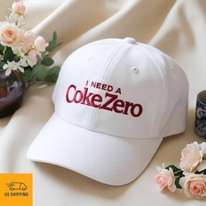 Op de afbeelding: Een witte baseballcap met de geborduurde tekst "I NEED A Coke Zero" in het rood. De cap heeft een gebogen klep en een klassiek zes-panelen ontwerp. De hoed wordt gepresenteerd op een zacht, lichtgekleurd oppervlak.