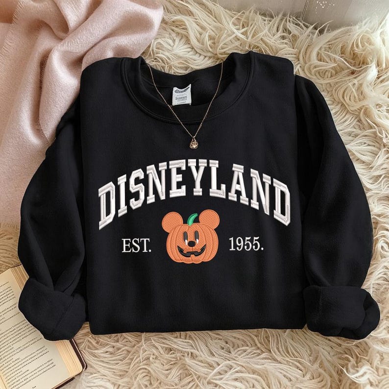 Może przedstawiać: Czarna bluza z okrągłym dekoltem z napisem "DISNEYLAND" w białych, łukowatych literach. Poniżej tekstu znajduje się pomarańczowa dynia z uszami Myszki Miki i napisem "EST. 1955."