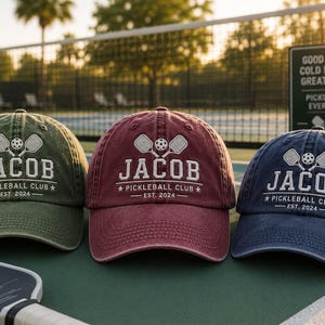 K&ouml;nnte beinhalten: Drei Baseballkappen in Olivgr&uuml;n, Burgunderrot und Marineblau, jede bestickt mit "JACOB PICKLEBALL CLUB EST. 2024" und Pickleball-Grafiken. Ein gelber Pickleball und ein Schl&auml;ger befinden sich im Vordergrund.