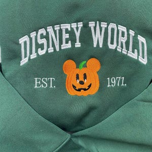 Może przedstawiać: Ciemnozielona bluza z haftowanym białym napisem "DISNEY WORLD". Poniżej głowa Myszki Miki w kształcie dyni z napisem "EST. 1971."