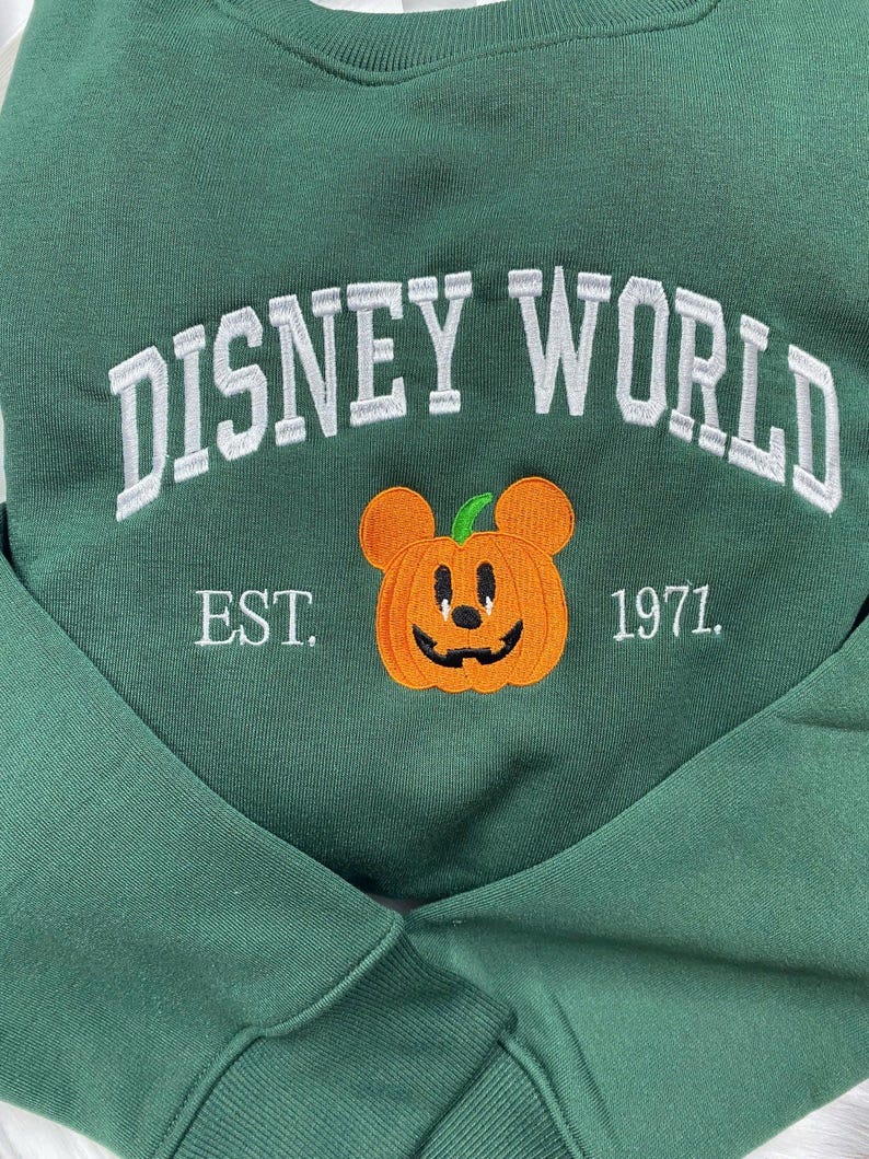 Może przedstawiać: Ciemnozielona bluza z haftowanym białym napisem "DISNEY WORLD". Poniżej głowa Myszki Miki w kształcie dyni z napisem "EST. 1971."