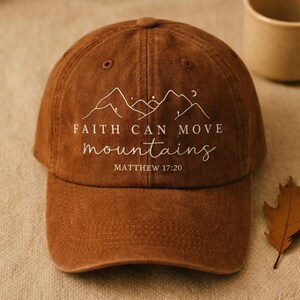May include: A brown baseball cap with the text "FAITH CAN MOVE mountains" and "MATTHEW 17:20" in white script. The cap features a mountain range design above the text. The cap is made of a soft, textured material.