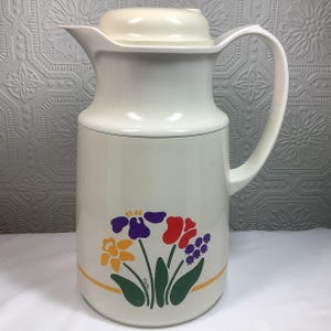May include: Cream-colored vintage thermos with a matching lid and handle. The thermos features a floral design with purple, red, yellow, and green accents. A thin orange stripe encircles the base of the floral design. The word "Tina" is printed on the floral design.