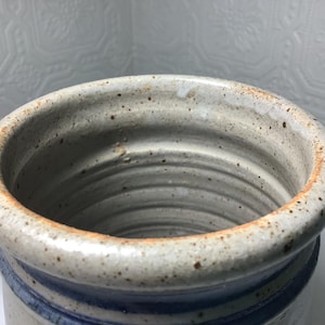 Puede incluir: Una vasija de cer&aacute;mica cil&iacute;ndrica con un esmalte blanquecino moteado y un borde grueso y redondeado. Las paredes interiores muestran estr&iacute;as horizontales del torno, mientras que una banda azul oscuro rodea la base exterior del recipiente.
