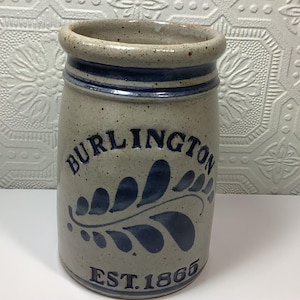 Puede incluir: Una vasija de gres cil&iacute;ndrica con un acabado gris claro moteado y un borde grueso. Presenta detalles pintados a mano en azul oscuro, incluyendo el texto BURLINGTON, un motivo de hoja y el texto EST. 1865 en la base.