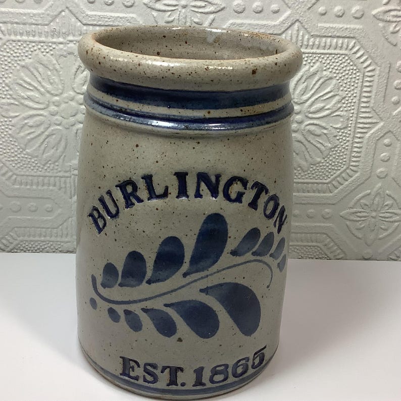 Puede incluir: Una vasija de gres cil&iacute;ndrica con un acabado gris claro moteado y un borde grueso. Presenta detalles pintados a mano en azul oscuro, incluyendo el texto BURLINGTON, un motivo de hoja y el texto EST. 1865 en la base.