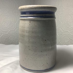 Puede incluir: Una vasija de cer&aacute;mica cil&iacute;ndrica con un esmalte moteado en color blanco roto y textura lisa. El recipiente est&aacute; acentuado por dos bandas azules horizontales distintas que rodean el borde superior y una banda azul en la base.