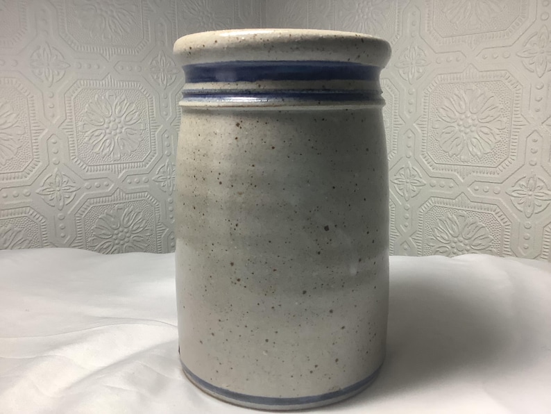Puede incluir: Una vasija de cer&aacute;mica cil&iacute;ndrica con un esmalte moteado en color blanco roto y textura lisa. El recipiente est&aacute; acentuado por dos bandas azules horizontales distintas que rodean el borde superior y una banda azul en la base.
