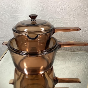 May include: A stack of three translucent amber-coloured glass saucepans with matching lids. Each saucepan features a long, integrated glass handle and a rounded body. The top saucepan includes a lid with a central knob, while the others are nested.