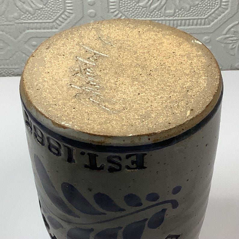 Puede incluir: Una vasija de gres cil&iacute;ndrica con acabado beige moteado y un patr&oacute;n de hojas pintado en azul oscuro. El texto EST. 1862 est&aacute; grabado en letras azul oscuro en la parte superior. La base no est&aacute; esmaltada y muestra una superficie de arcilla.