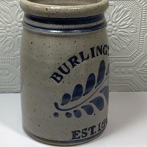 Puede incluir: Una vasija de gres cil&iacute;ndrica con un acabado beige moteado y un borde azul oscuro. El frente presenta el texto en azul oscuro BURLINGTON y EST. 1800, acompa&ntilde;ado de un dise&ntilde;o de hoja pintado en azul oscuro. El recipiente tiene una superficie esmaltada.