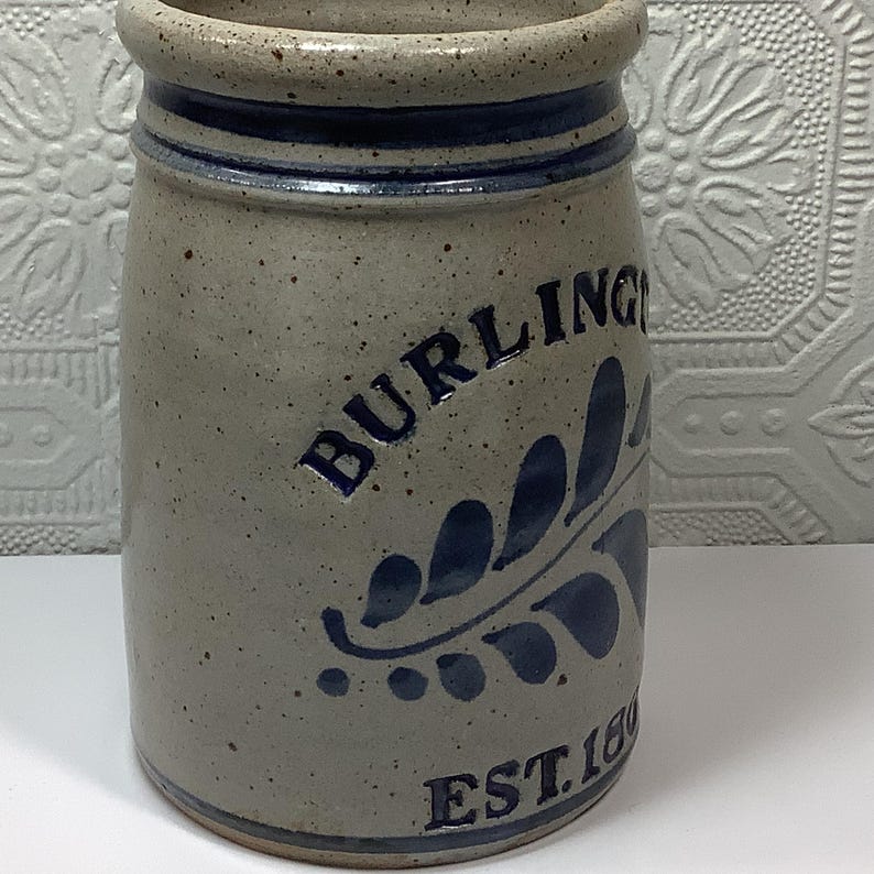 Puede incluir: Una vasija de gres cil&iacute;ndrica con un acabado beige moteado y un borde azul oscuro. El frente presenta el texto en azul oscuro BURLINGTON y EST. 1800, acompa&ntilde;ado de un dise&ntilde;o de hoja pintado en azul oscuro. El recipiente tiene una superficie esmaltada.