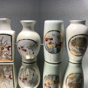 May include: Four white ceramic vases with gold detailing and floral patterns. The vases are in various shapes, including rectangular and rounded. Each vase has a unique illustration of flowers, butterflies, and birds.